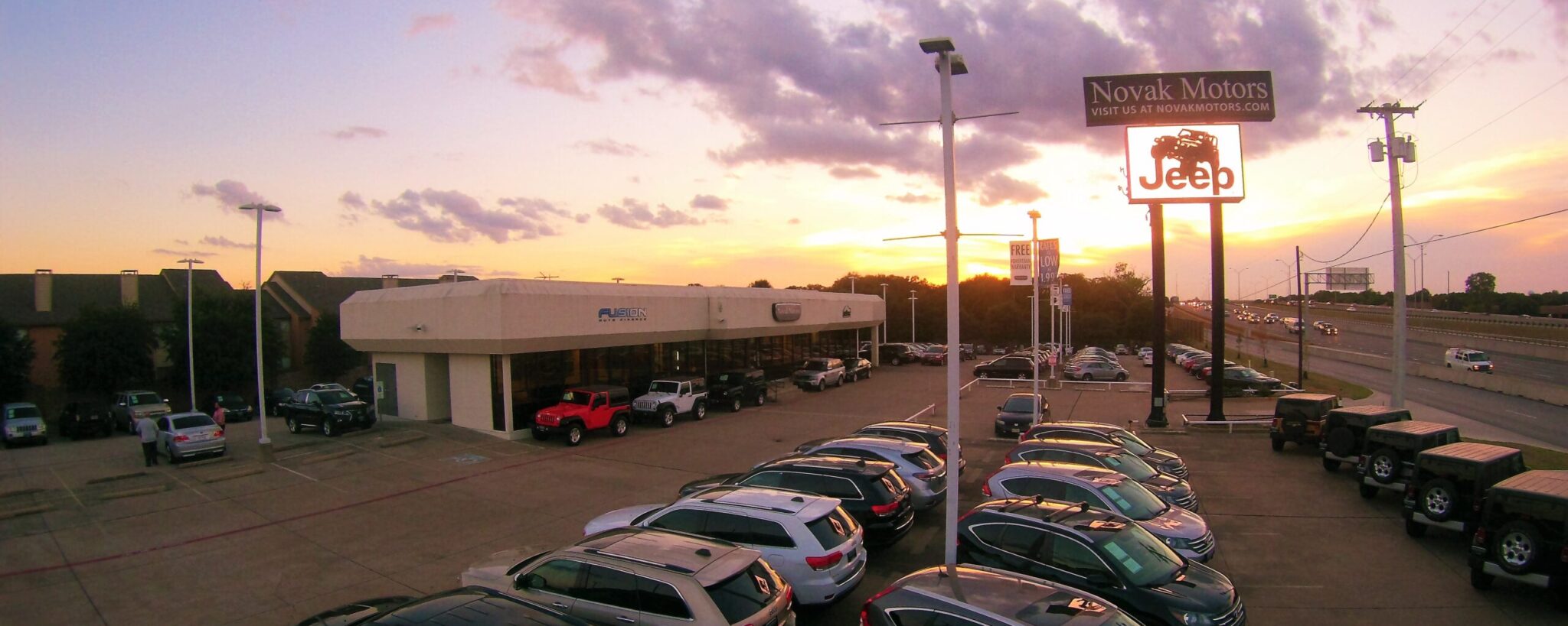 Sunset view of Novak Motors dealership in Bedford, Texas, showcasing parked vehicles and a Jeep sign, celebrating Fusion Auto Finance’s 2012 milestone of 20,000 vehicles leased through CU Xpress Lease.